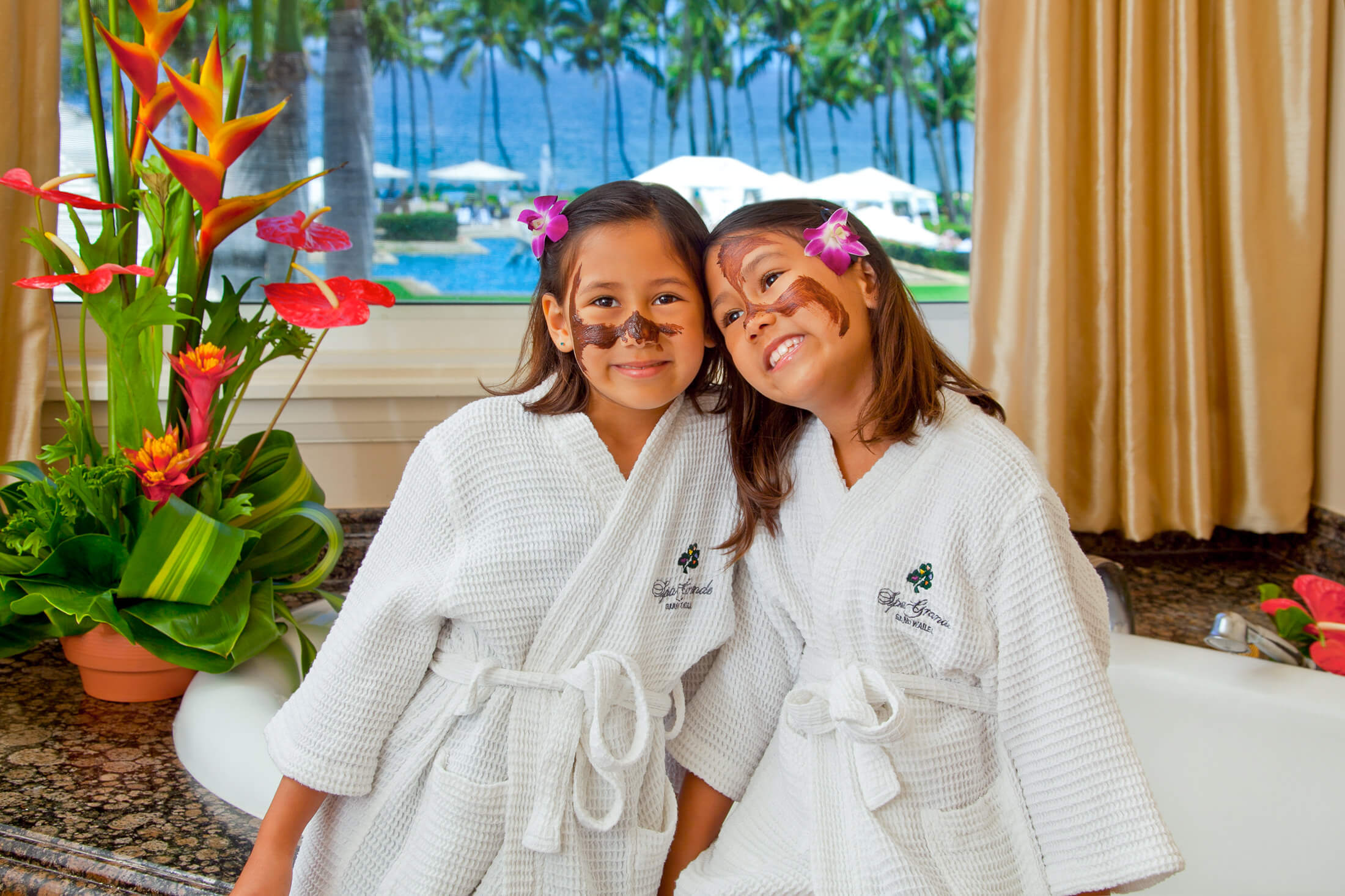 two kids wearing spa grande robes with mud masks on their cheeks sit on edge of tub