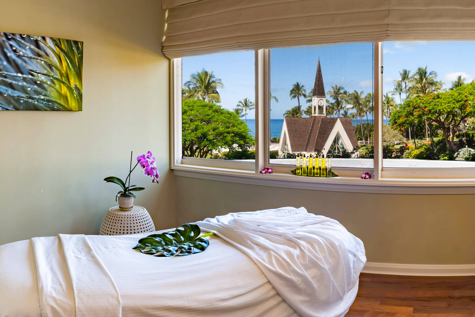 a massage table covered in a white blanket topped with flowers with a view of the Grand Wailea chapel out the window