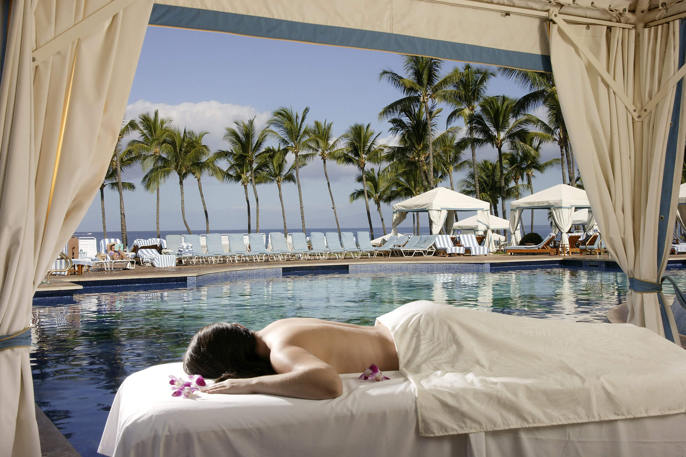 a woman lays on her stomach on a massage table with curtains open to the Grand Waila pool and cabanas