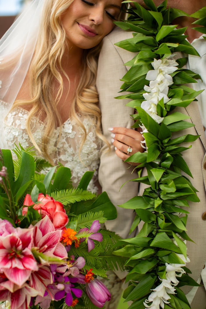 A bride holding a pink flower bouquet holding the arm of her groom and resting her head on his shoulder