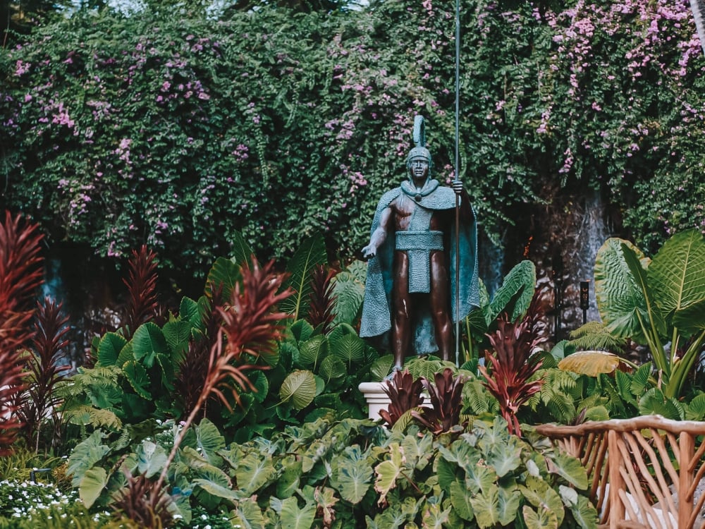 a statue of warrior stands amongst green foliage in front of a forest of trees