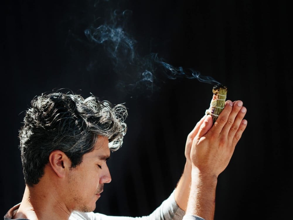 side profile of a man sitting crosslegged on the ground holds his hands up while smoke trails over his head