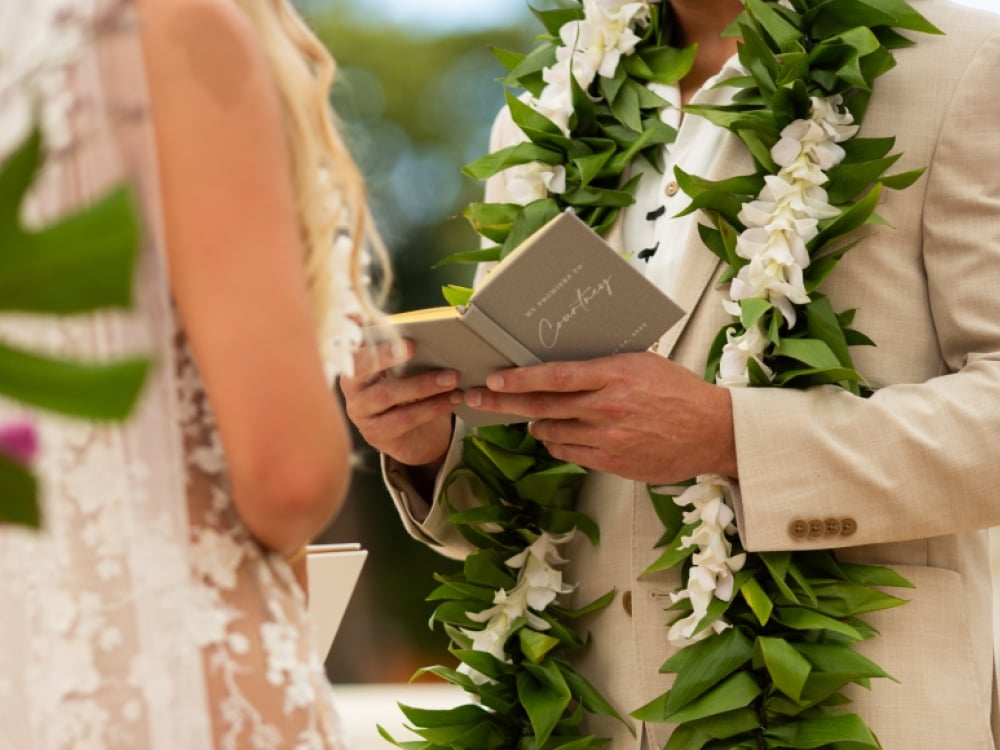 A groom with a lei reads vows from a book at an outdoor wedding