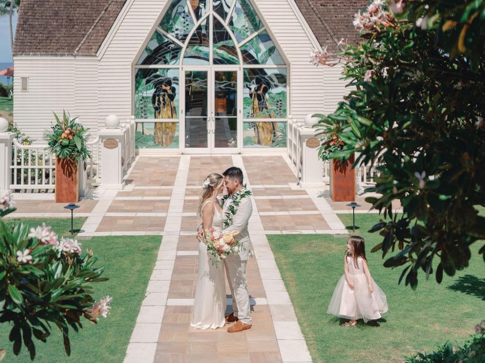 A bride and groom standing in front of a chapel with a little flower girl next to them