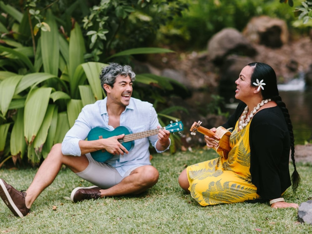 A man seated in the grass with a woman, receiving a ukelele lesson