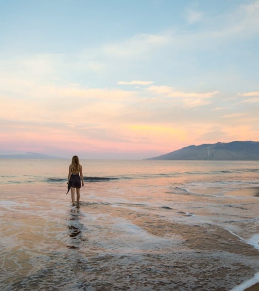 a woman walks toward the ocean on the beach with mountains in the background while the sun sets