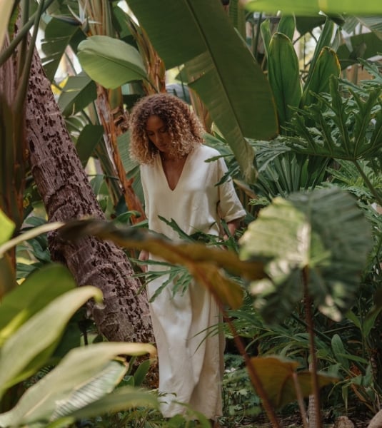 A woman walks through a forested garden area with large leafy green trees and and bushes