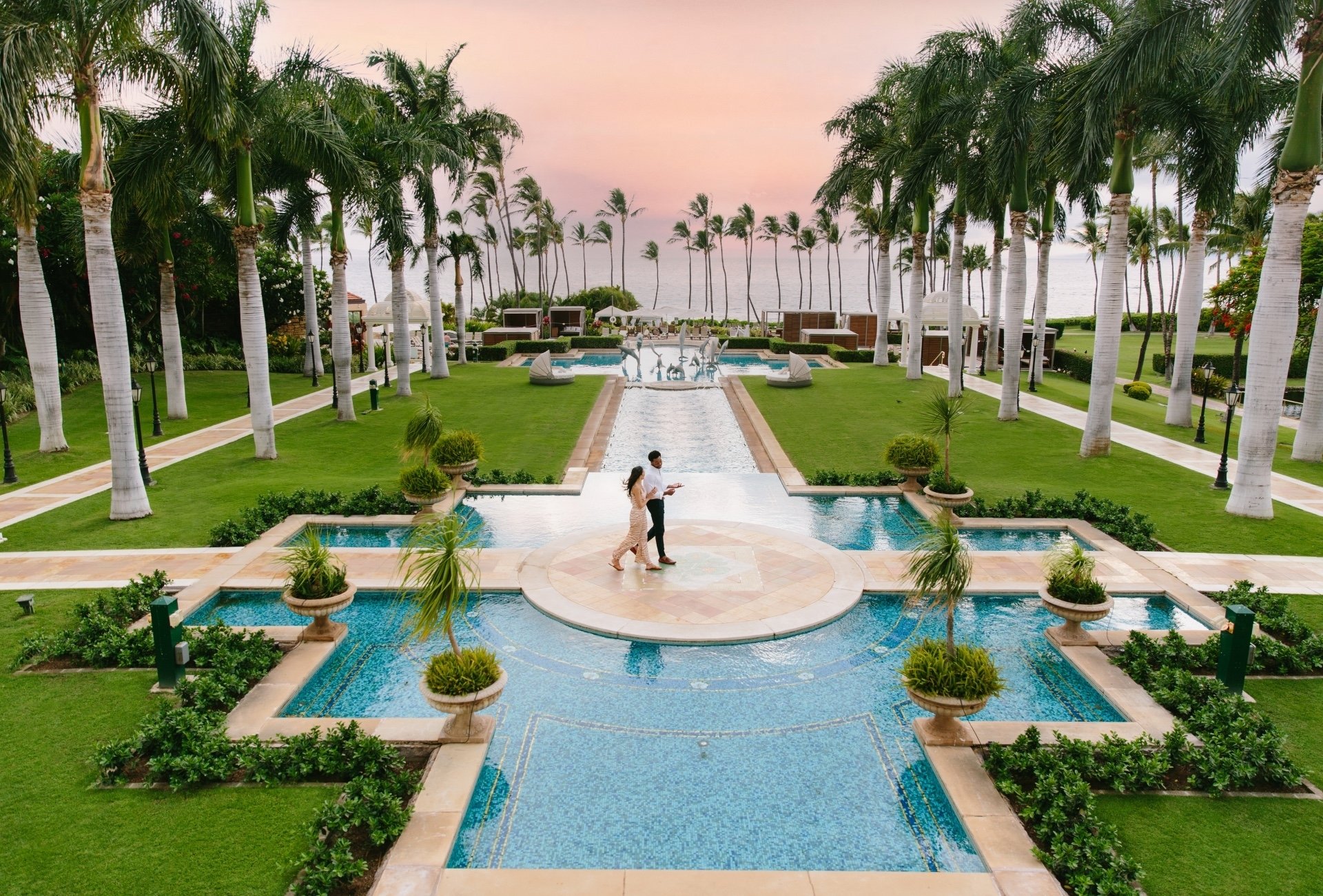 A couple walks across a bridge spanning a long pool running along the 